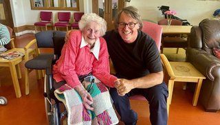 A woman sitting in a wheelchair with a blanket on her lap. A man is sitting beside her in a chair.