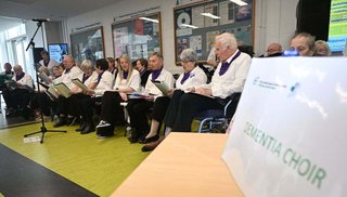 A group of people sitting. They are holding notebooks and singing. A sign reads 'Dementia Choir'.