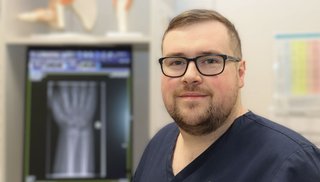 A healthcare worker in a clinical setting wearing scrubs and glasses