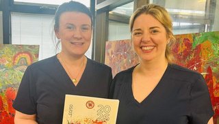 Two women wearing scrubs smiling into camera. There is an art display in the background.