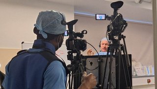 A man behind a camera recording a healthcare worker in a hospital