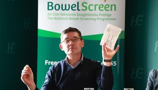 A man holding a bowel screening at home testing kit. There's a pull up banner in the background that reads "BowelScreen".