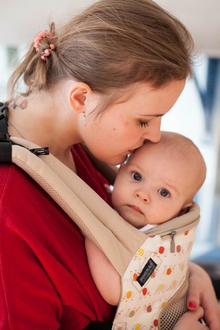 A mother kissing a baby who is positioned closely to her chest in a baby carrier sling