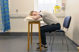 Physio sitting on chair leaning forward resting his elbows on a table. He is using a pillow for comfort.