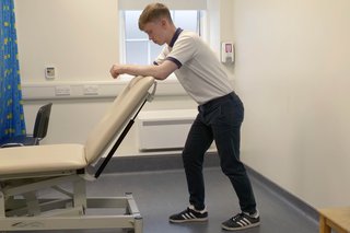 Physio is standing up leaning forward, resting his elbows on the back of a chair