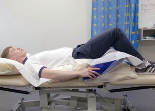 Physio lying on his back with head flat and knees bent. Pillow is under hips and knees so that hips are higher than the level of his chest.