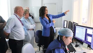 A small group of people in a room in a clinical setting, looking through large windows into another room. There are a number of computer screens on a desk facing the windows and a healthcare worker sits in front of one of them, speaking into a microphone.