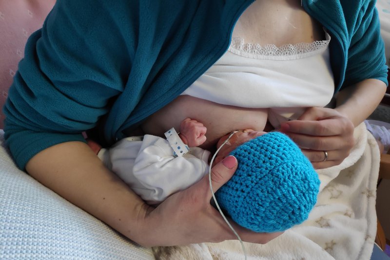 Woman holding her premature baby while breastfeeding. The baby&#x27;s legs are under the arm and around the woman&#x27;s back.