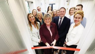 A group of people smiling. A woman is cutting a ribbon with a pair of scissors