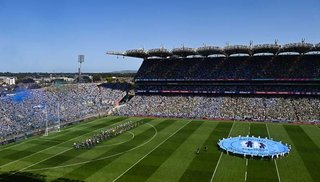 A large, wide view of the inside of Croke Park GAA stadium, with a marching band and both teams lined up on the right and a group of people on the right holding a large, unfurled circular banner showing the 'Dementia: Understand together' logo.