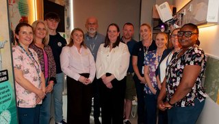 Eleven people stand together in a clinical room. They are all smiling and some are wearing healthcare clothes.