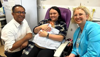 Mother smiling while holding newborn baby while sitting in purple hospital chair. There is a man to her left and a woman to her right.