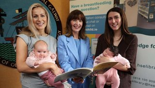 Three women standing. Two women are holding babies and one woman is holding a brochure.
