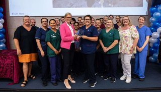 A large group of healthcare staff standing on a stage and smiling. Two women in the centre are shaking hands and holding a plaque.