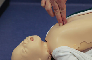 First aider demonstrating chest compressions on an infant CPR dummy. They have two fingers in the middle of the chest and are pushing inwards.