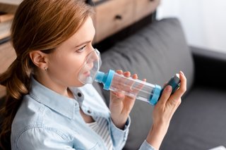 A person using an inhaler with a spacer. The mask of the spacer is covering their mouth and nose. They are pressing down the canister of the inhaler.