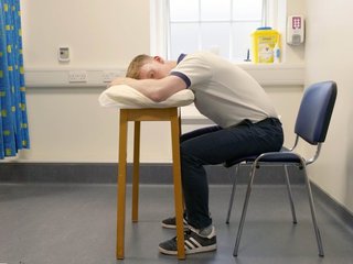 Image of a boy sitting on a chair, leaning forward and resting his head sideways on a pillow.