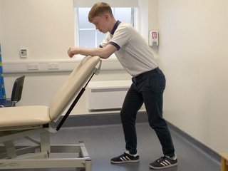 Image of a boy leaning forward and resting his elbows on the back of a chair, with 1 foot in front of the other and bent knees