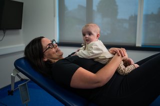 A woman lying propped up at a slight angle on an adjustable bed with a baby on her chest.