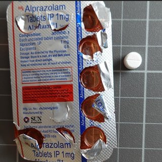 The back of a counterfeit alprazolam blister pack next to a small white tablet. Several of the tablets are missing to show the orange colour of the front of the packet.