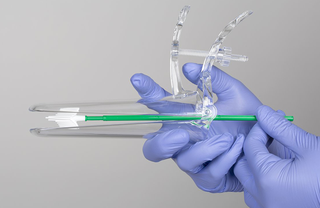 A pair of blue-gloved hands holding a plastic transparent speculum which has a small white brush at its centre